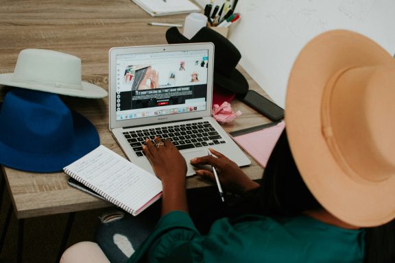 Person working on a laptop surrounded by hats and a notebook.