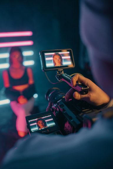 A person is being filmed with a camera, featuring neon lights in the background.