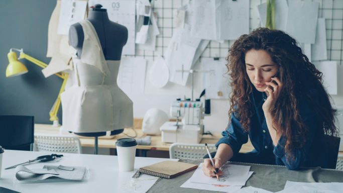 A fashion designer sketches plans while talking on the phone, surrounded by tools and patterns.